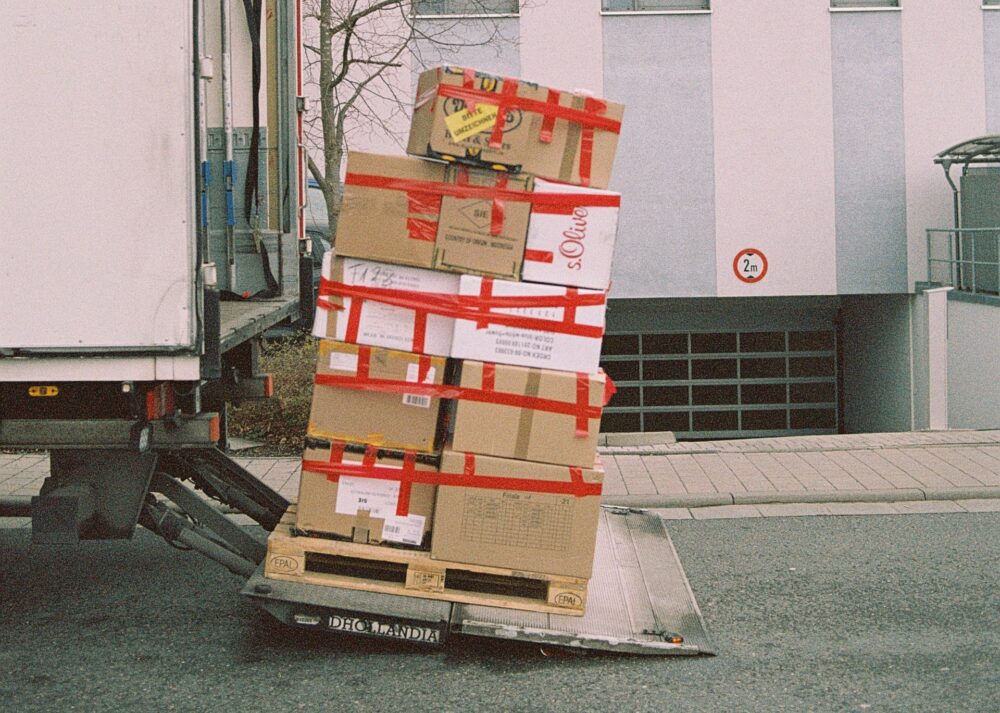 A stack of cardboard boxes secured with red tape is being lowered on a hydraulic lift from a delivery truck. The truck is parked on a street next to a sidewalk, with a building in the background. Some boxes are tilting slightly to one side.