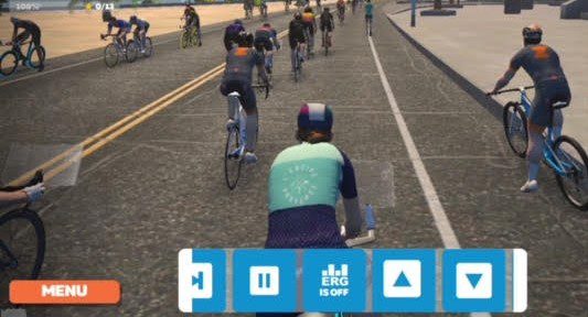 A virtual cycling competition is displayed on a screen. The view shows multiple cyclists on a road, with game controls visible at the bottom of the screen. The cyclists are in different outfits, and the environment includes buildings and a clear sky.