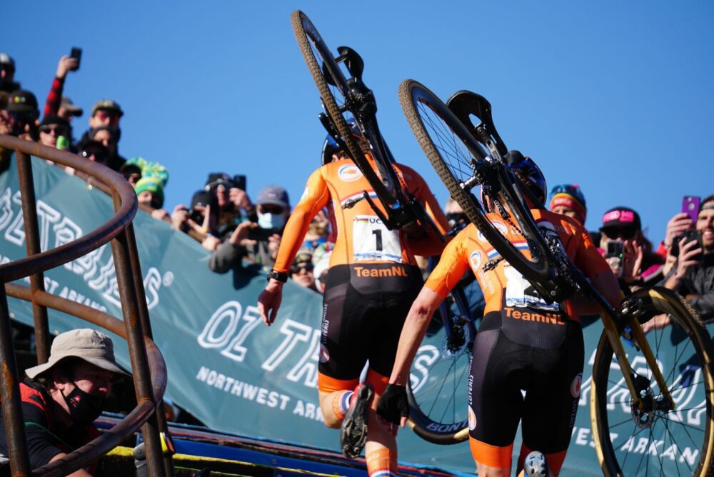 Two cyclists in orange jerseys from Team NL are carrying their bicycles up a staircase during a race. They are surrounded by spectators, some of whom are wearing masks. The background features a banner with text and a clear blue sky.