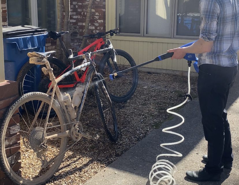A person wearing a plaid shirt and black pants is using an electric pressure washer to clean a dirt-covered bicycle. Another clean bicycle and a blue recycling bin are nearby against a brick wall. The scene takes place outside a building on a sunny day.