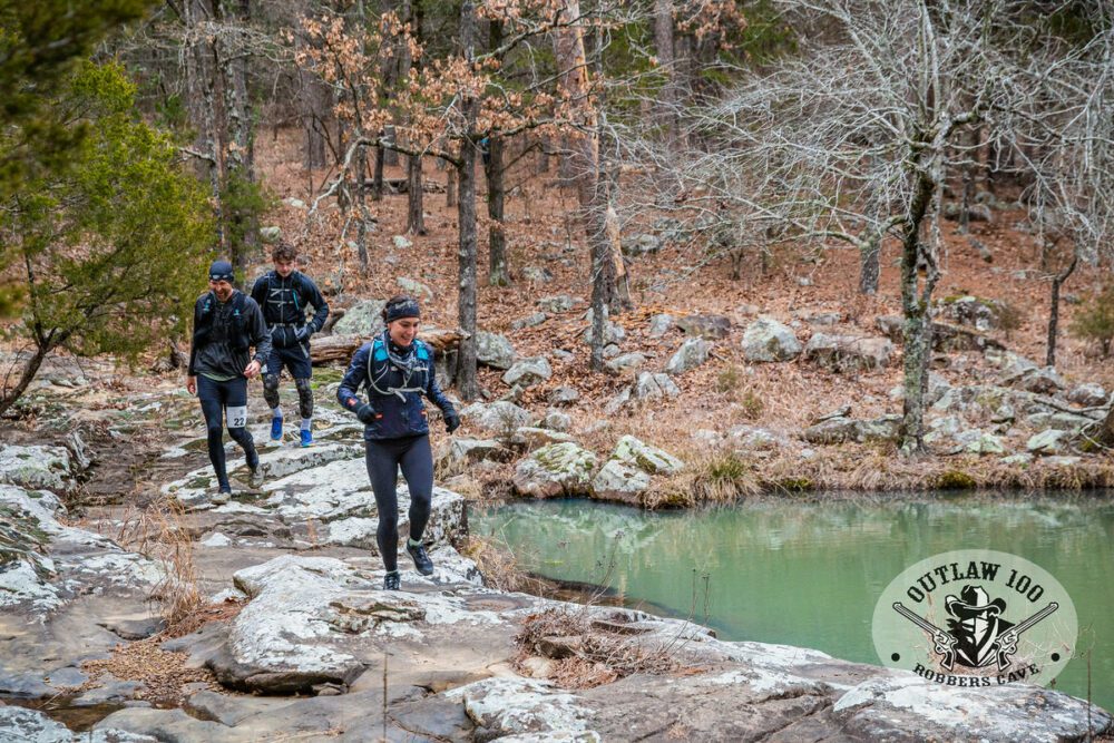 Three trail runners wearing cold weather gear are running along a rocky path beside a small body of water in a forested area. The trees are mostly bare, and the ground is covered in fallen leaves and rocks. A logo in the bottom right corner says "Outlaw 100.