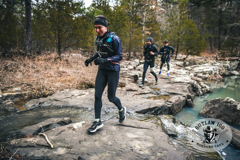 A woman leads two men running on rocky terrain beside a clear stream in a forested area during an outdoor adventure race, with the event logo "outlaw 100 - robbers cave" overlaid on the image.
