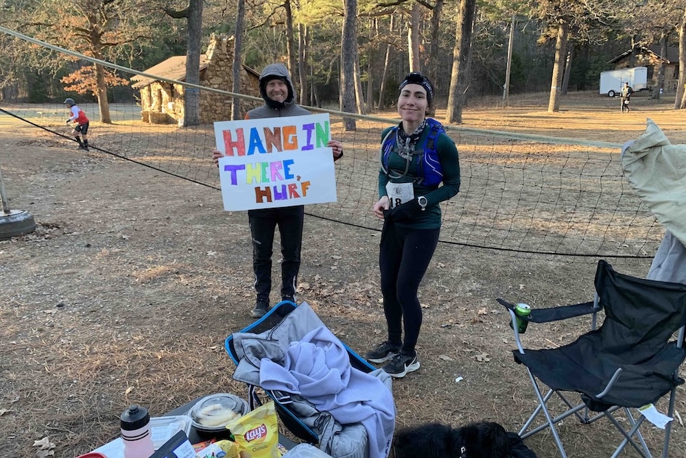 Two individuals at an outdoor cycling training event; one is holding a sign that reads "hang in there hurf," while the other, possibly a participant, is smiling and appears to be wearing a race