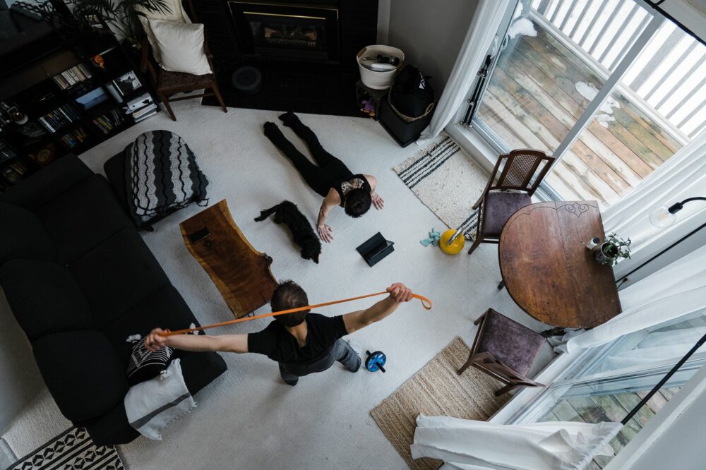 Overhead view of a living room with two people and a cat. One person stretches with a resistance band near a laptop, while another lies face down with their arms extended. The room features a fireplace, bookshelf, coffee table, and sliding glass door.