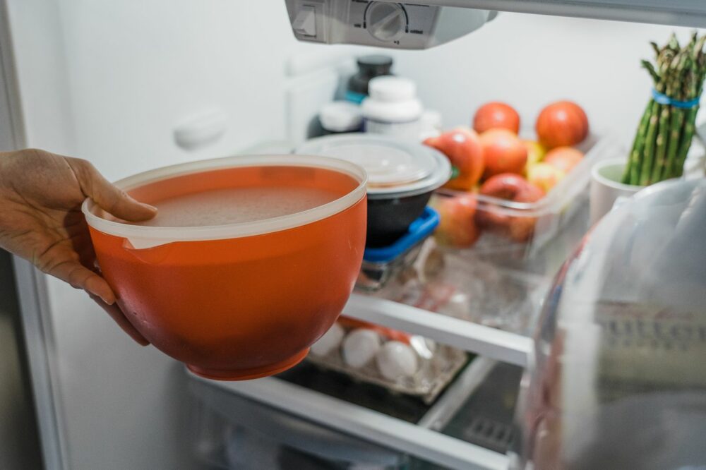 A person places a sealed red bowl into a refrigerator. Inside the refrigerator, there are containers of food, apples, a bunch of asparagus, eggs, and several other items.