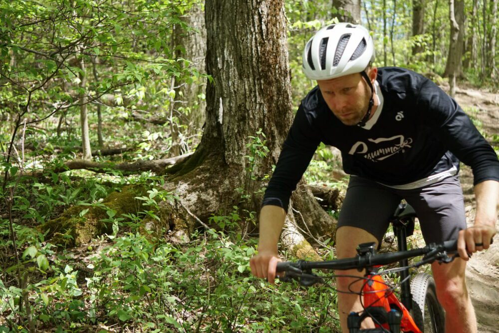 A cyclist wearing a helmet, black shirt, and shorts rides a mountain bike through a wooded trail. The large trees and greenery suggest a forested area.