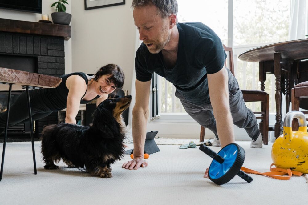 A man and a woman are doing push-ups in a living room. The man is close to a small black and brown dog looking at him. Exercise equipment like a kettlebell and an ab roller are on the floor. The woman is smiling in the background.