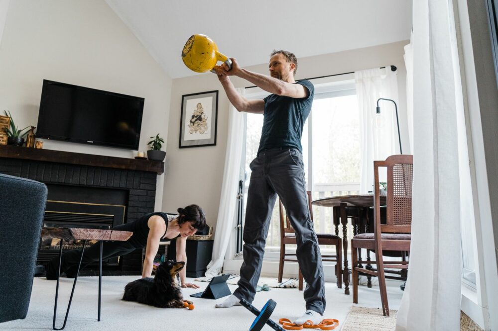 A man is exercising with a yellow kettlebell in a living room, while a woman is in a plank position nearby. A black and brown dog is resting on the floor between them. The room is well-lit with natural light coming through large windows.