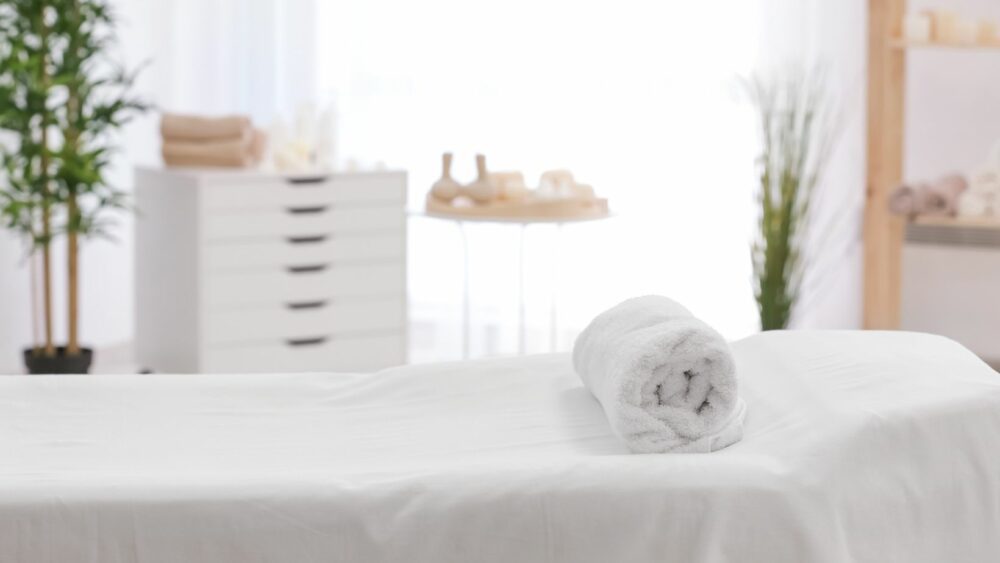 A rolled white towel lies on a neatly made massage table in a bright, airy spa room, with soft lighting and organized shelves in the background.