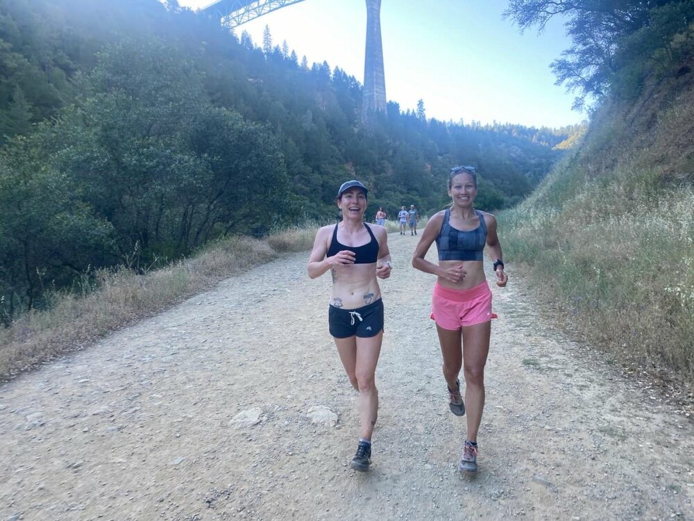 Two women are running on a dirt trail through a hilly, forested area. One wears a black sports bra and shorts, while the other wears a gray sports bra and pink shorts. A tall bridge is visible in the background, spanning a deep valley. Other runners are in the distance.