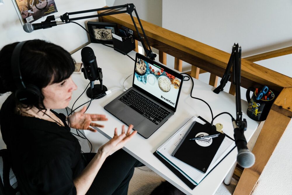 A person wearing headphones speaks into a microphone, gesturing with their hands. They sit at a desk with a laptop open to a website, a notebook, and a potted plant. Two microphone arms are attached to the desk. The setting appears to be a recording or podcast space.