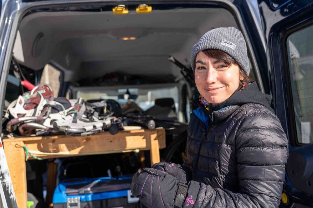A person wearing a gray beanie and black puffer jacket stands in front of a blue vehicle. The back of the vehicle is open, revealing various equipment, including climbing or hiking gear. The person is smiling and wearing black gloves.