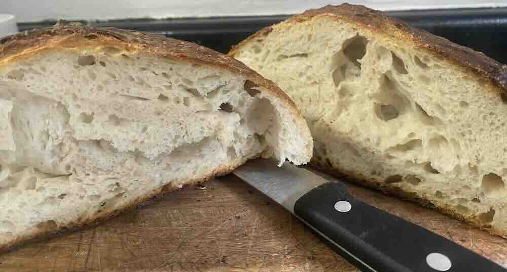 A loaf of crusty bread cut in half, revealing its airy and slightly dense interior with several large holes. A knife with a black handle is placed between the two halves on a wooden cutting board.