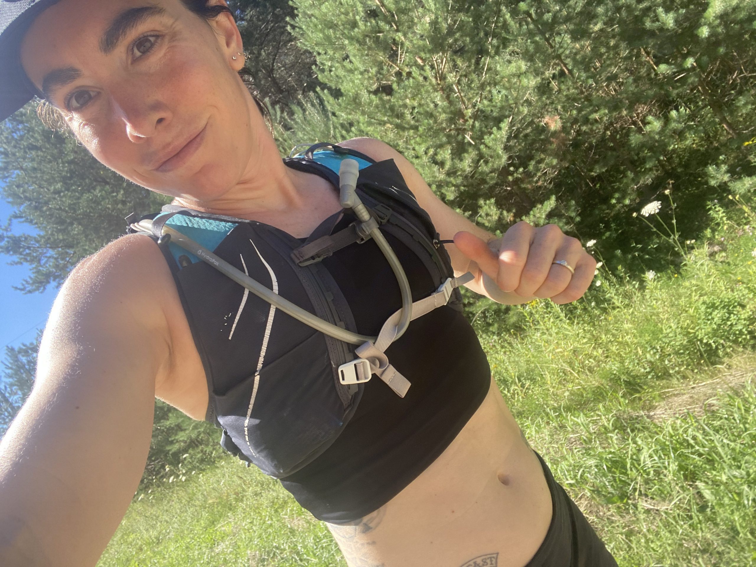 A person wearing a sleeveless sports top and hydration pack is outside in a grassy and forested area. They are taking a selfie, and the image is upside down. Sunlight is casting shadows, and the sky is clear and blue.