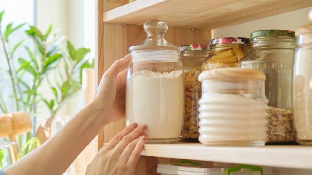 A person is placing a glass jar of white flour onto a wooden shelf. The shelf also contains other jars filled with pasta, oats, and sugar, all with wooden or sealed lids. A green plant is visible in the background.