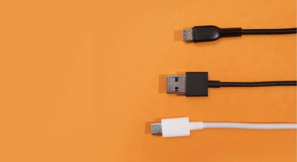 Three different electronic cables are aligned horizontally on an orange background. From top to bottom: a black micro USB cable, a black USB Type-A cable, and a white USB Type-C cable.