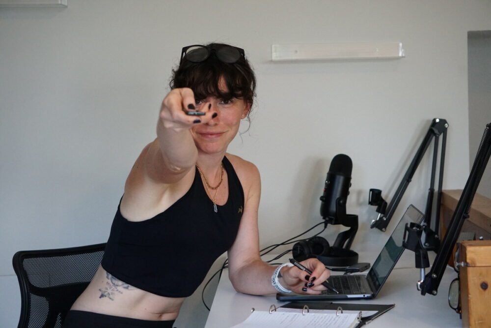 A person wearing a black tank top, sunglasses on their head, and black nail polish, is sitting at a desk with a laptop, microphones, headphones, and a notebook. They are pointing directly towards the camera.