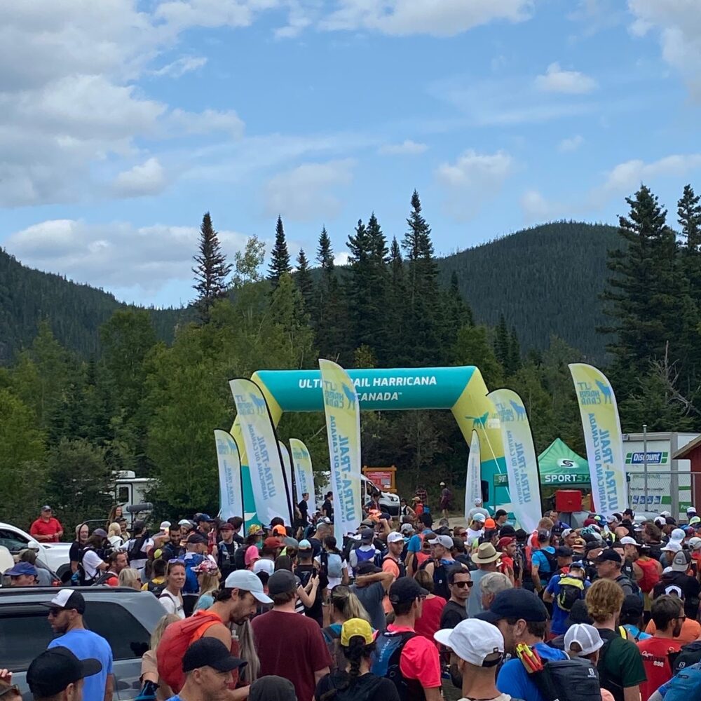 A large group of people gather at the starting area of the Ultra-Trail Harricana in Canada. The crowd is near a blue and green inflatable archway, surrounded by flags and trees, under a partly cloudy sky.