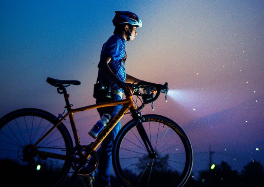 A cyclist stands with a road bike, wearing a helmet and cycling attire. The bike's front light is turned on, illuminating small particles in the air. The background shows a dusky sky transitioning from blue to purple.