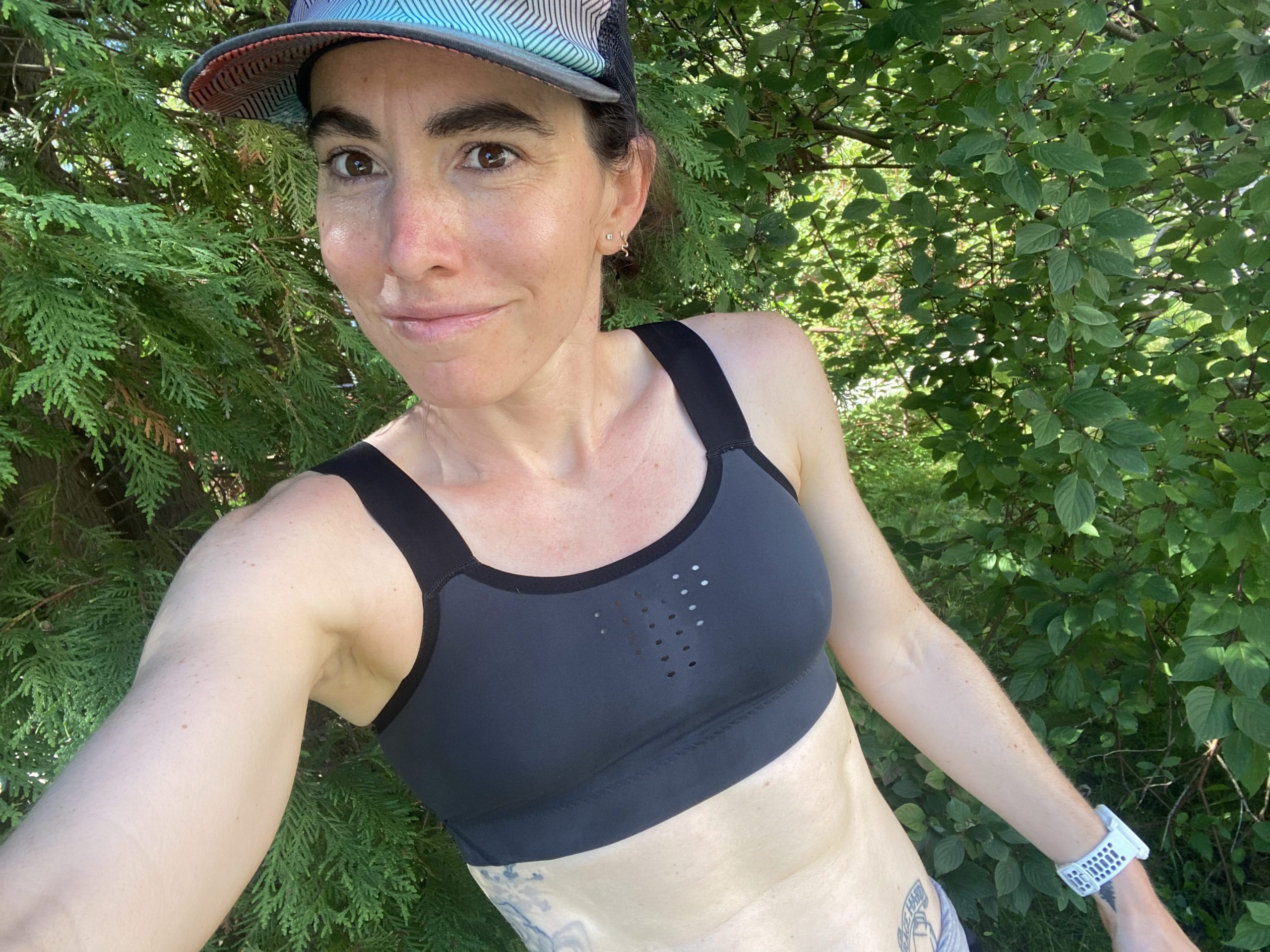 A person wearing a sports bra and a cap is posing outdoors amidst greenery. The individual has tattoos on their arms and appears to be engaging in a physical activity or exercise routine.