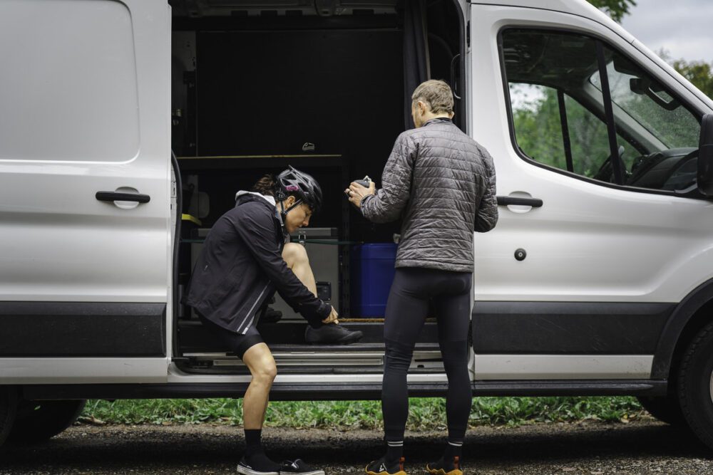 Two people in cycling attire stand beside an open van. One person is seated inside the van, adjusting their shoe, while the other stands outside, appearing engaged in conversation and holding gear. The van is parked on a gravel surface with greenery visible in the background.