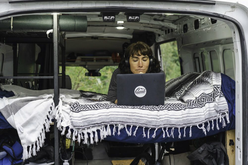 A person sits on a bed inside a van intently working on a laptop. The van is retrofitted with bedding and storage, and the person is wearing large headphones. The back doors are open, revealing an outdoor setting with greenery in the background.