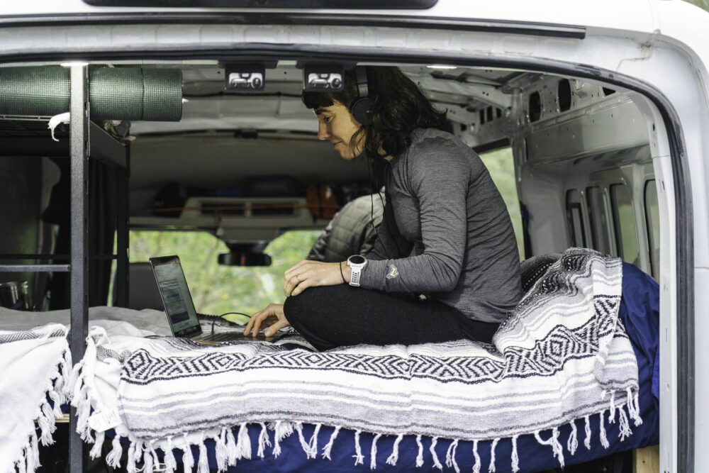 A person is sitting cross-legged on a blanket-covered bed inside a van, wearing headphones and using a laptop. The van’s back doors are open, revealing a green outdoor background. Camping and gear can be seen in the interior of the van.