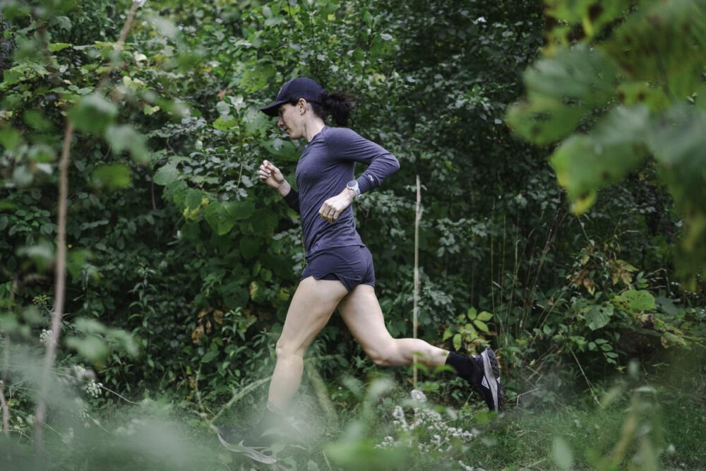 A person wearing a dark long-sleeve shirt, shorts, black cap, and running shoes runs through a densely forested area. The individual is captured mid-stride, surrounded by green foliage and natural vegetation.