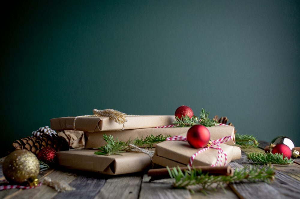 Several brown paper-wrapped gifts are arranged on a wooden surface with festive decorations, including red and gold baubles, pine cones, and green pine branches, against a dark green background.