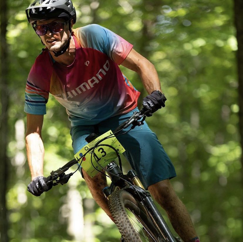 A cyclist wearing a multicolored Shimano jersey, blue shorts, and protective gear, including a helmet and sunglasses, is riding a mountain bike on a forest trail. The bike has a race number "3" attached to the front. The background is lush and green.