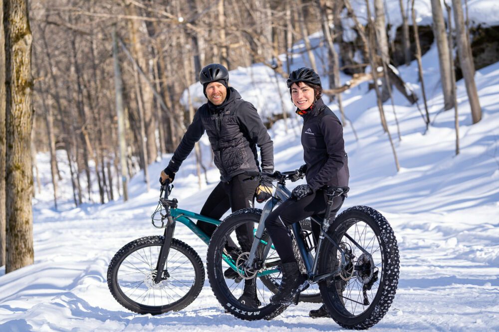Two people wearing helmets and winter cycling gear are riding fat bikes on a snowy trail in a forest. The sun is shining, and leafless trees are visible in the background. Both riders are looking at the camera and smiling.