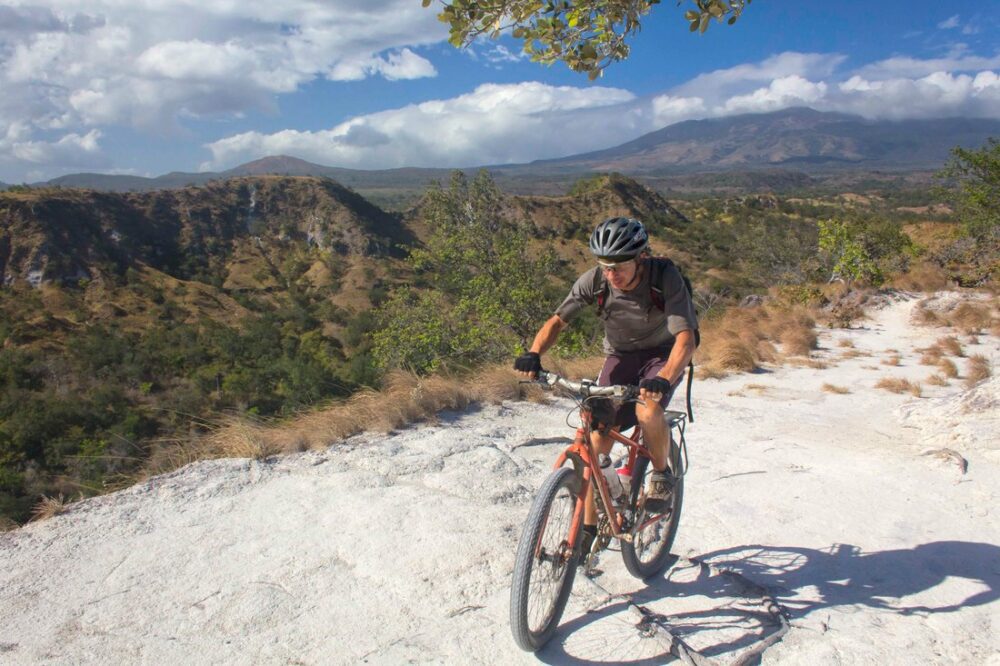 A person wearing a helmet and outdoor gear rides a mountain bike on a rocky trail. The background features a landscape with hills, sparse vegetation, and distant mountains under a partly cloudy sky.