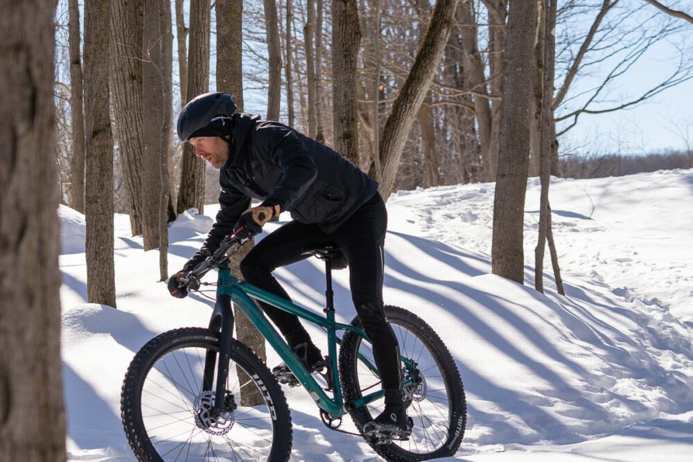 A person wearing a black helmet and black outdoor clothing is riding a blue mountain bike on a snowy trail through a forest. The trees are leafless, suggesting it is winter. The sun is shining, casting shadows on the snow.