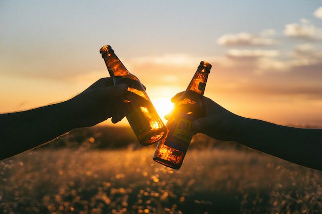 Two hands clink beer bottles together against a sunset backdrop in an open field. The golden light of the sunset illuminates the bottles, creating a warm and relaxed atmosphere.