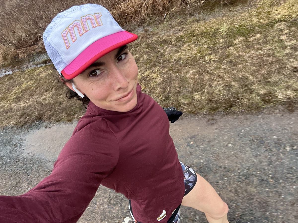 A person wearing a maroon long-sleeve shirt, patterned shorts, and a white cap with pink text is captured in a selfie while walking or jogging on a dirt path. The background shows dry grass along the path. The image is taken from an overhead angle.
