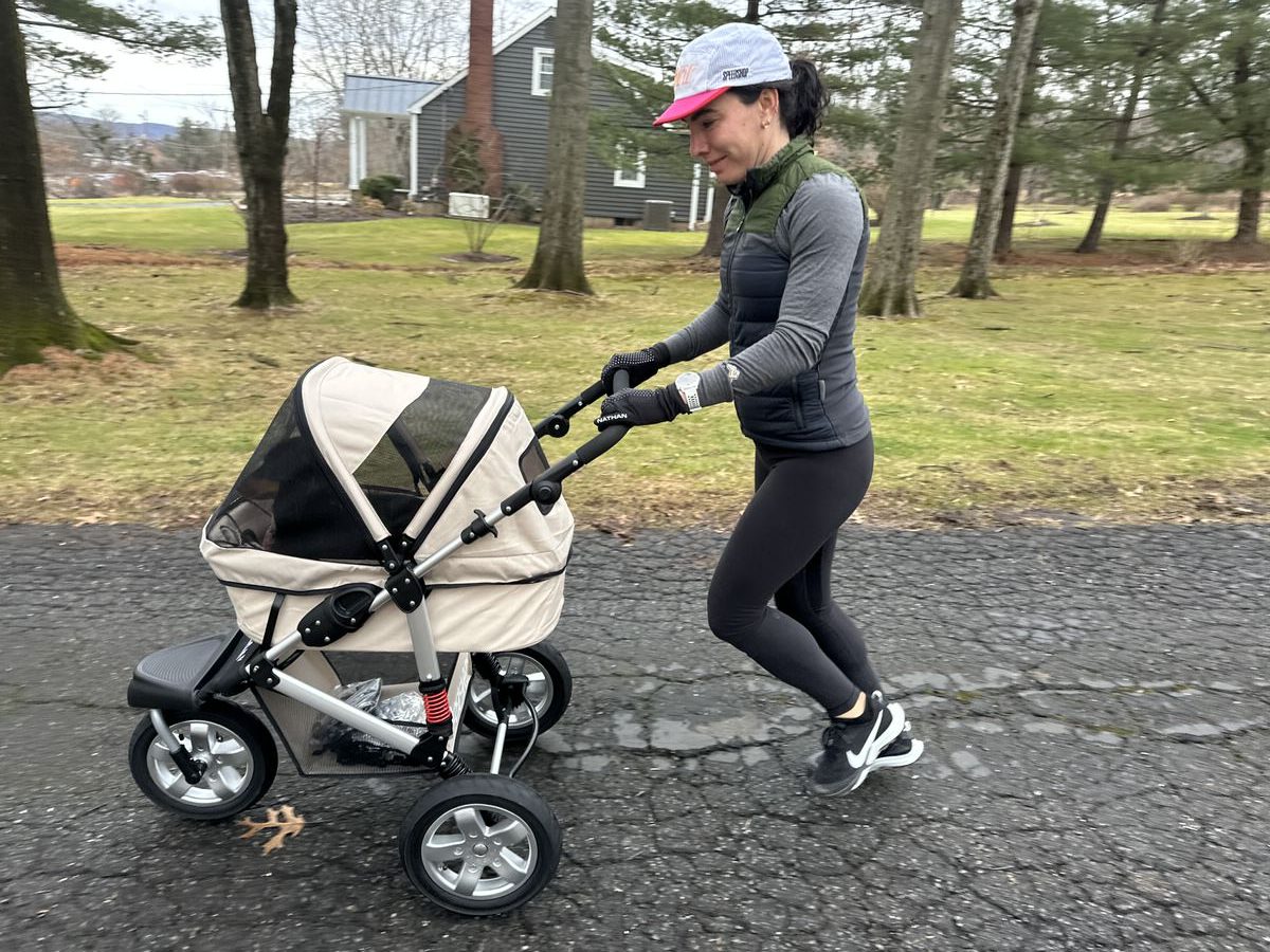 A woman jogs on a paved path while pushing a baby stroller. She wears a gray long-sleeve shirt, black leggings, white sneakers, and a white visor. The background includes grass, trees, and houses. The weather appears overcast.