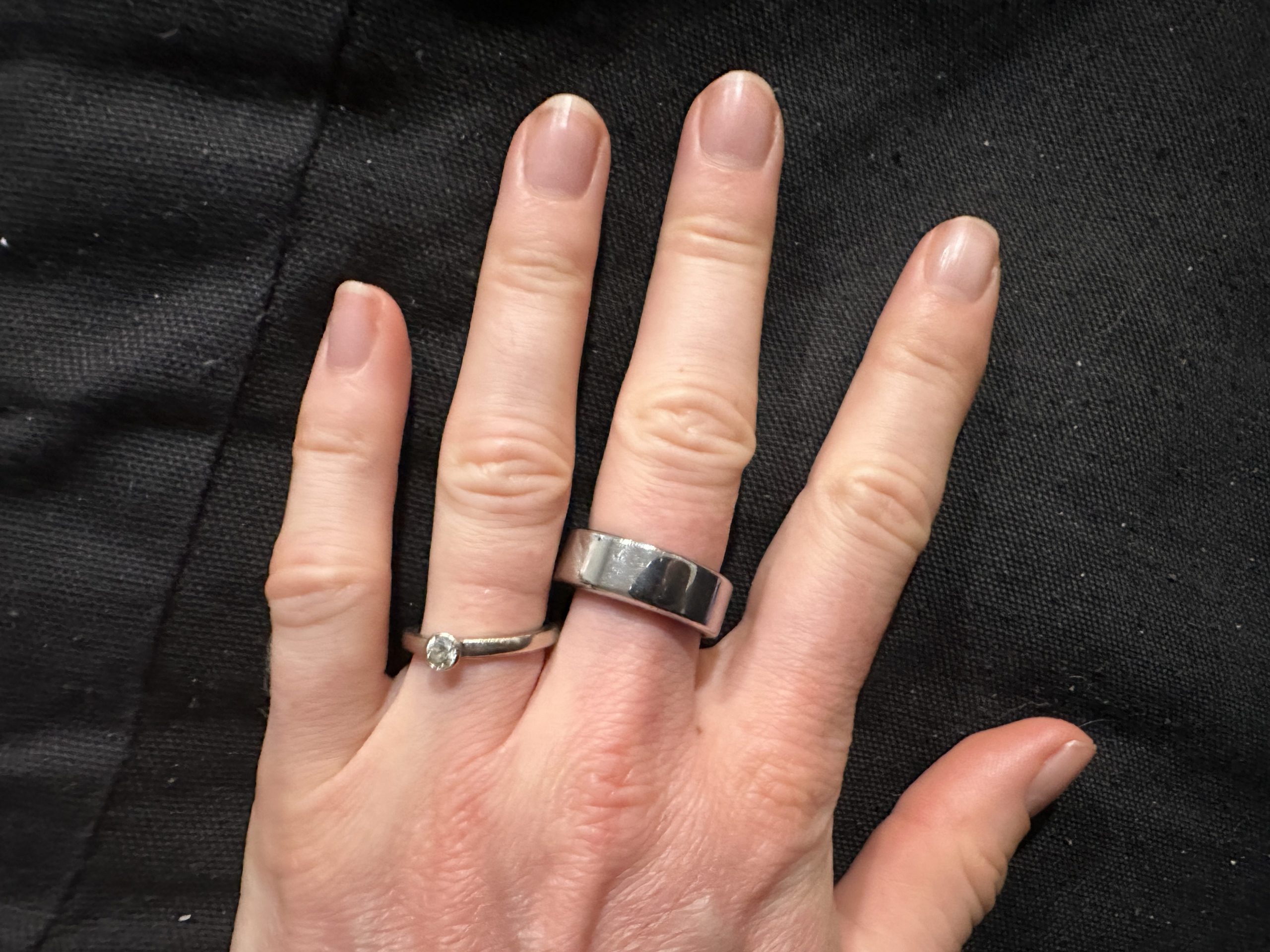 A hand with a light skin tone is shown against a dark fabric background. The hand is wearing two rings: a silver band on the middle finger and a silver ring with a clear stone on the ring finger. The fingernails are short and neatly trimmed.