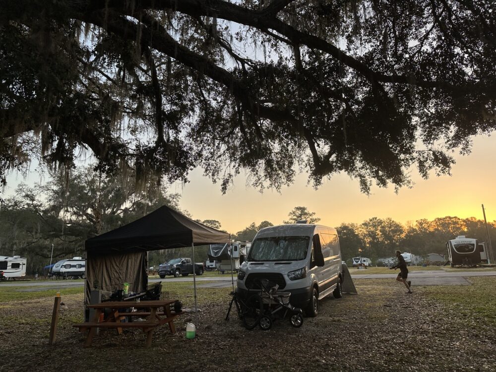 A campsite at sunset with a parked white camper van next to a canopy shelter, a picnic table with items on top, and a stroller in front. Several other RVs are visible in the background under the silhouette of large, mossy tree branches.