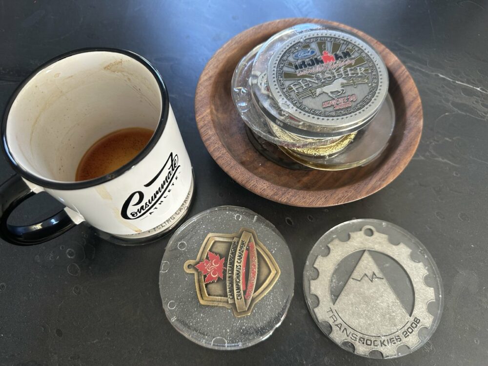 A coffee cup with a small amount of coffee sits on a black surface. Next to it, there is a wooden bowl filled with various medals. Two more medals are laid out in front of the bowl, featuring different designs.