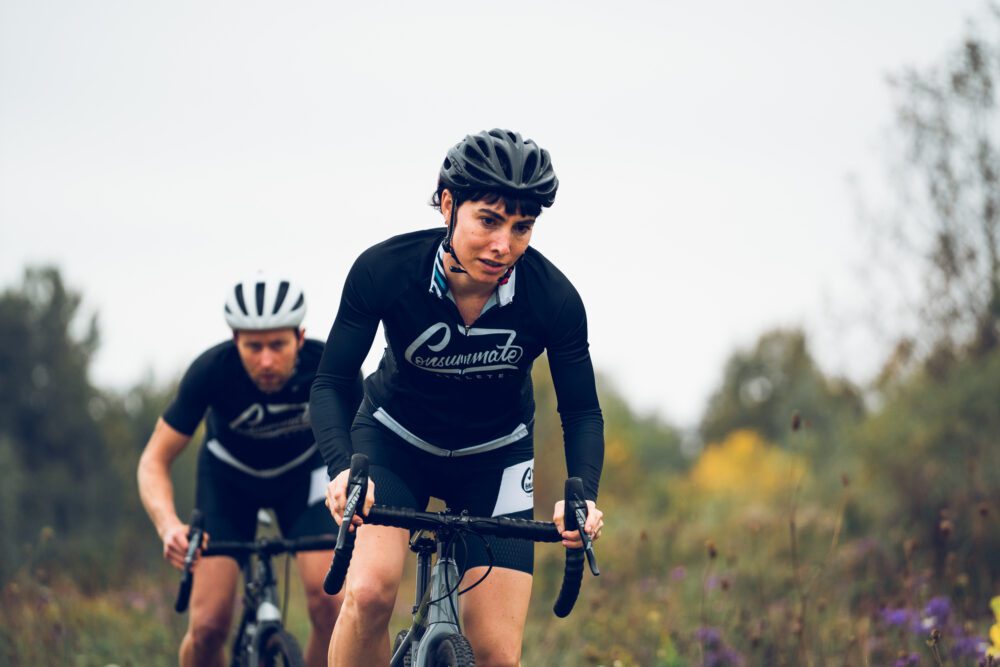 Two cyclists wearing black jerseys and helmets ride on a path with greenery in the background. Both appear focused and are riding road bikes. The scene is outdoors, and the sky is overcast.