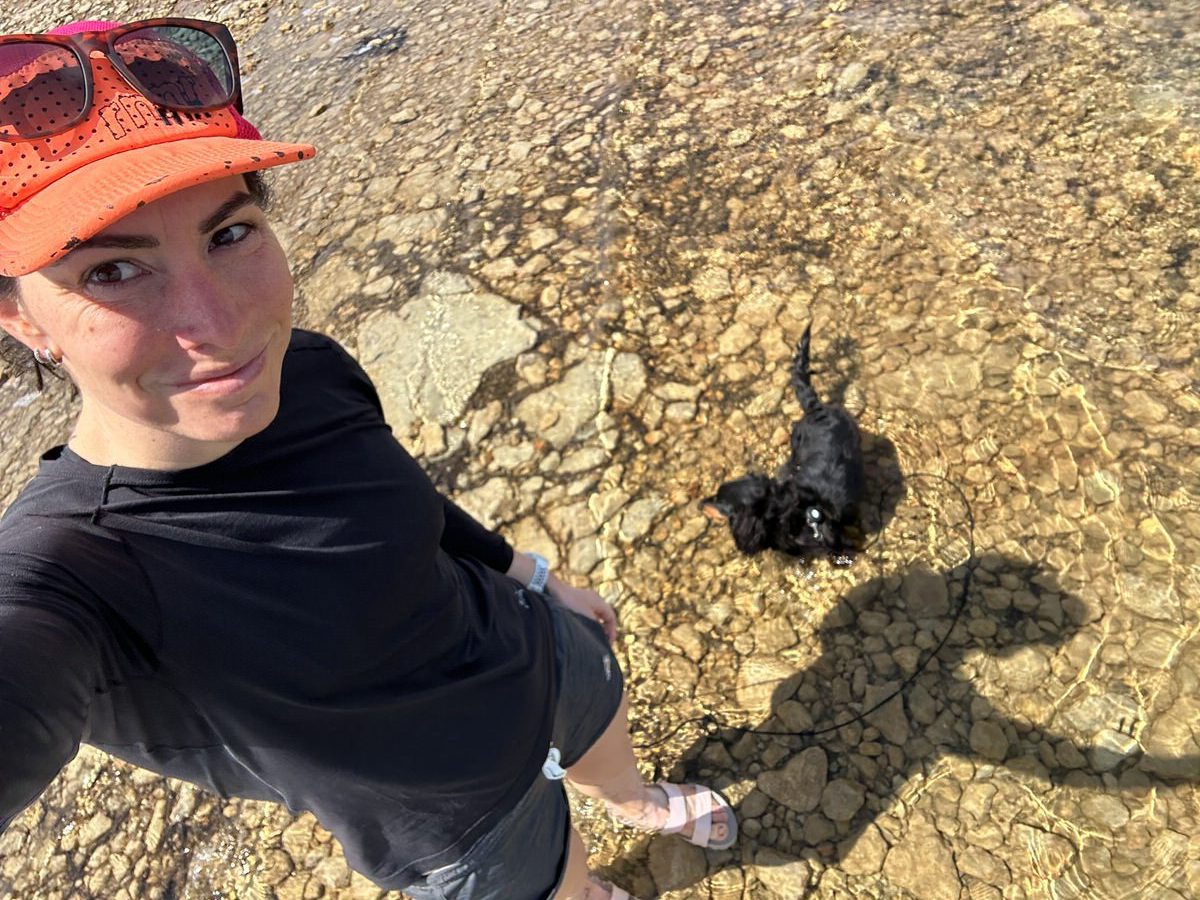 A person wearing a black shirt, orange cap, and sunglasses taking a selfie at a rocky beach. Their shadow and that of a small black dog are visible on the rocky ground. The person is standing in very shallow, clear water.