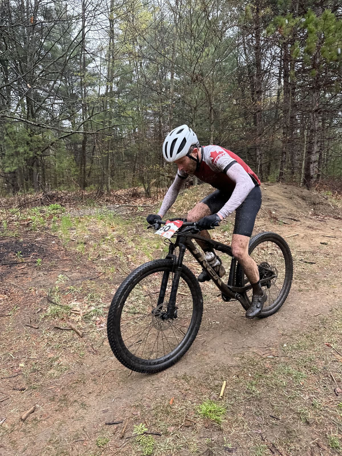 A cyclist wearing a white helmet, red and white clothing, and riding a black mountain bike navigates a dirt trail within a forested area. The ground is damp and scattered with leaves and twigs, suggesting recent rain.
