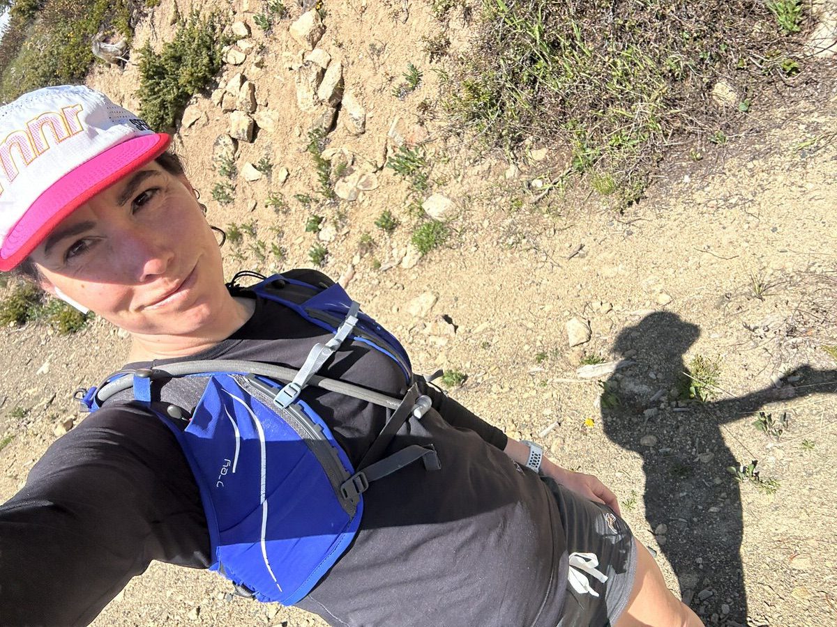 A person wearing a blue hydration backpack, black long-sleeve shirt, and a white and pink cap is outdoors on a rocky trail. The person appears to be on a hike or run, and a shadow is cast on the ground beside them.