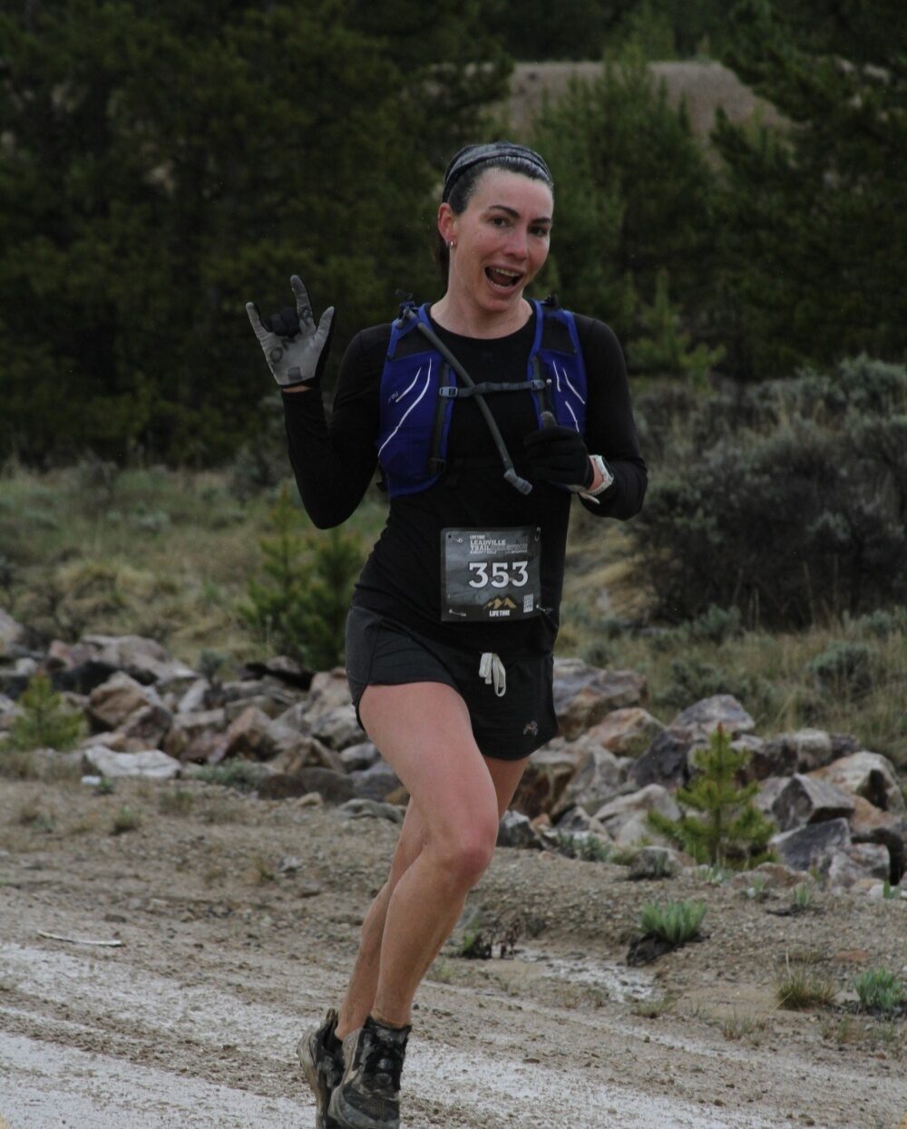A person wearing a black outfit and a blue hydration pack is running on a dirt path with vegetation and rocks around. They are holding up their left hand in a gesture with the index finger, pinky finger, and thumb extended. They are wearing a race bib numbered 353.