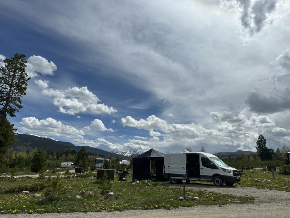 A white van with its side door open is parked near a small black canopy in a grassy field under a partly cloudy sky. There are trees and mountains in the distance and a few other vehicles and tents scattered around.