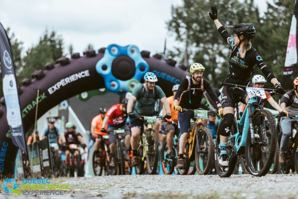 A group of cyclists is seen participating in the Québec Singletrack Experience event. The riders are at the starting line, passing under an archway that reads "EXPERIENCE," while one cyclist raises their right arm in an enthusiastic gesture.