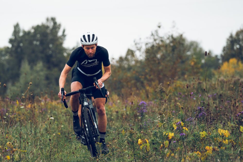 A cyclist wearing a black outfit and white helmet rides a bicycle on a grassy trail surrounded by wildflowers and greenery. Trees are visible in the background under an overcast sky.