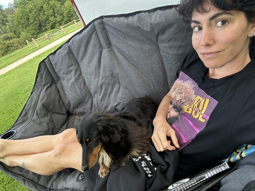 A person with dark hair and a black shirt featuring a graphic design is sitting on a reclining chair outdoors. A long-haired dachshund is lying on their lap. There is greenery and a wooden fence visible in the background.