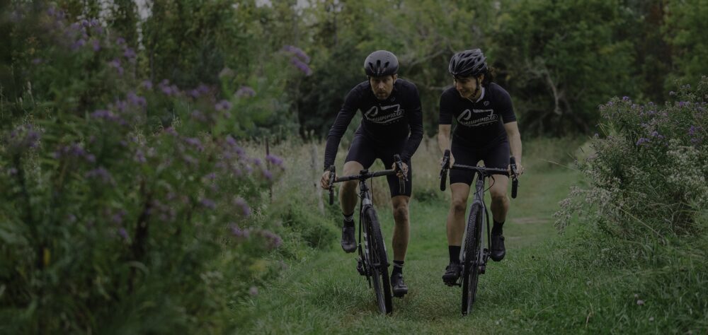 Two cyclists ride on a grassy trail surrounded by lush greenery and purple wildflowers. Both are wearing black cycling outfits and helmets, with the cyclist on the left positioned slightly ahead of the one on the right.