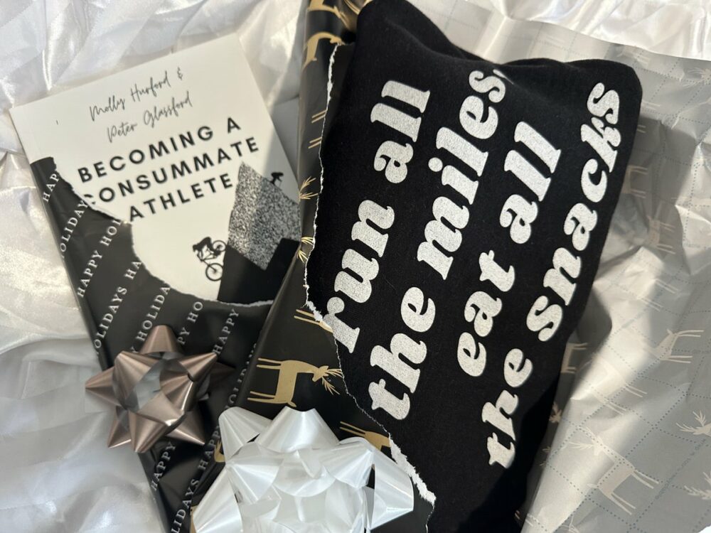 A gift-wrapped package is shown. Inside, there is a book titled "Becoming a Consummate Athlete" and a black item of clothing with white text that reads "run all the miles, eat all the snacks." Additionally, there are silver and white bows on the package.